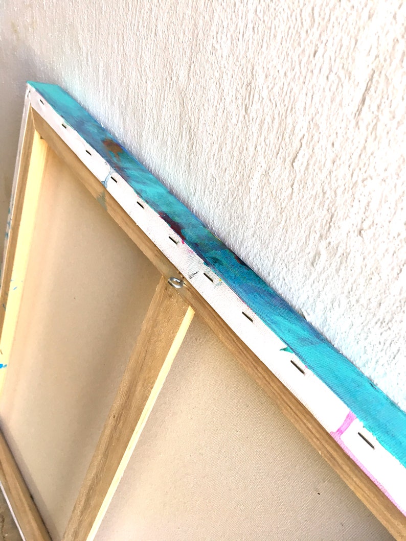 May include: Close-up of the back of a stretched canvas painting. The canvas is wrapped around a wooden frame and secured with staples. The painting features a blue, green, and pink abstract design.