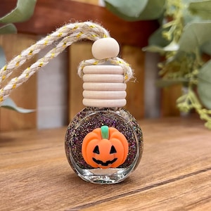 May include: A small, round glass car air freshener bottle filled with dark purple and gold glitter. The bottle features a bright orange jack-o'-lantern pumpkin charm on the front, a light wood ribbed cap, and a beige rope for hanging.
