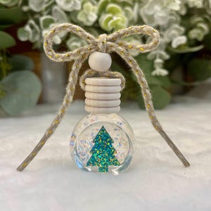 May include: A clear glass car air freshener bottle featuring a teal glitter Christmas tree inside, filled with iridescent glitter. The bottle has a ribbed wooden cap, a round wooden bead, and a beige braided cord tied into a decorative bow.