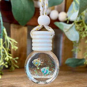 May include: A clear, round glass car air freshener bottle with a light wood cap and a braided rope hanger. Inside the bottle, two small fish figures are suspended in liquid with iridescent glitter particles that catch the light.