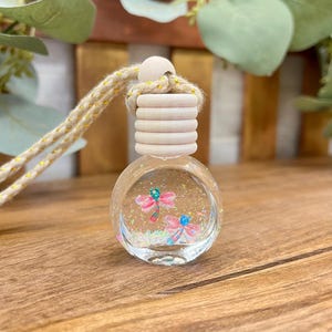 May include: A small, round, clear glass bottle filled with liquid, iridescent glitter, and two pink dragonfly charms. The bottle features a light-colored, ribbed wooden cap and is suspended by a thick, beige, braided cord with gold threading.