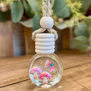 May include: A round glass car air freshener bottle filled with liquid and iridescent glitter. Inside the bottle are two small pink mushrooms with white spots and a pink dragonfly. The bottle has a white wooden cap and a braided rope for hanging.