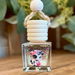 May include: A square glass car air freshener bottle featuring a cartoon cow graphic with a pink bow and heart. The bottle is filled with iridescent glitter and topped with a ribbed wooden cap and a braided rope hanger with a round wooden bead.