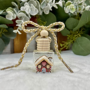 May include: A small square glass bottle filled with clear liquid, topped with a light wood cap and a braided beige and gold cord tied in a bow. The front of the bottle features a decorative gingerbread house charm with a red heart and candy cane trim.