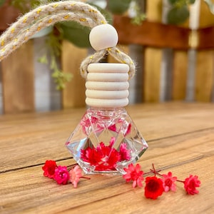 May include: A diamond-shaped glass car air freshener bottle filled with clear liquid and bright pink dried flowers. The bottle features a ribbed wooden cap, a round wooden bead, and a thick braided jute hanging cord with gold metallic thread.