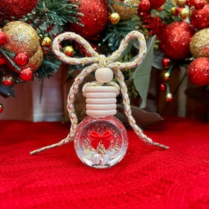 May include: A clear glass car air freshener bottle with a wooden cap and a beige braided rope tied in a bow. Inside the round bottle is a red bow charm and iridescent glitter. The bottle features a small wooden bead above the cap.