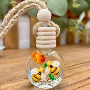 May include: A small, round, clear glass bottle filled with liquid, containing two miniature yellow and black bumblebee figurines and a small orange flower. The bottle is topped with a ribbed wooden cap and hangs from a braided tan rope.