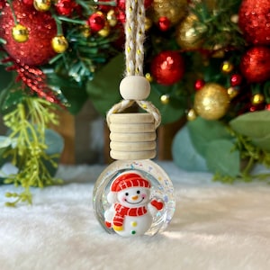 May include: A clear glass spherical car air freshener bottle featuring a small snowman figurine inside. The snowman wears a red hat and scarf. The bottle has a light-colored wooden cap and a braided jute hanging cord with a wooden bead accent.