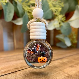 May include: A round glass car air freshener bottle filled with dark glittery liquid. The front features a small bat and a black cat sitting on an orange jack-o'-lantern. It has a light wooden cap and a braided rope hanger with gold metallic threads.