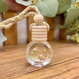 May include: A small, round glass bottle hanging from a braided beige rope with gold threading. The bottle features a light wood cap and contains liquid with iridescent glitter and two miniature orange and blue fish figurines inside.