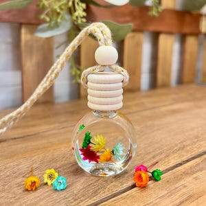 May include: A round glass car air freshener bottle filled with clear liquid and small colorful dried flowers in shades of yellow, orange, pink, and blue. The bottle has a light-colored wooden cap and a thick, braided beige cord with gold threading.
