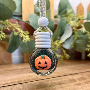 May include: A small, round glass car air freshener bottle filled with dark, iridescent glitter. The bottle features a white wooden cap and a hanging cord. An orange, smiling jack-o'-lantern pumpkin charm is attached to the front of the glass.