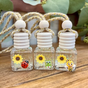 May include: Three square glass bottles with light wood caps and hanging ropes. Each bottle contains clear liquid with glitter and features a yellow sunflower charm paired with a ladybug, a green frog, or a yellow bee character charm.