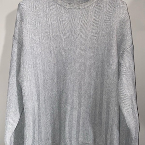 May include: A light gray crew neck sweater with a subtle herringbone pattern. The sweater has long sleeves and a ribbed hem and cuffs. The brand name "haggare" is visible on the black hanger.