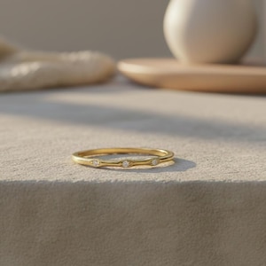 Puede incluir: Un delicado anillo de oro con tres pequeñas piedras preciosas transparentes. El anillo tiene un acabado pulido y está colocado sobre una superficie neutra. El diseño del anillo es simple y elegante.