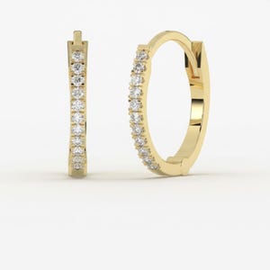 Może przedstawiać: Para złotych kolczyków w kształcie obręczy. Każdy kolczyk jest inkrustowany małymi, przezroczystymi kamieniami szlachetnymi wzdłuż zewnętrznej krzywizny. Kolczyki mają prosty, elegancki design, odpowiedni do codziennego użytku lub na specjalne okazje.