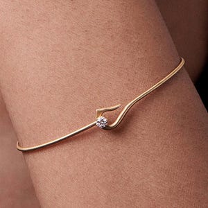 May include: A delicate gold-tone bracelet with a small, sparkling gemstone at the center. The bracelet features a unique, curved design. The gemstone is round and appears to be a diamond or a similar clear stone. The bracelet is worn on the arm.