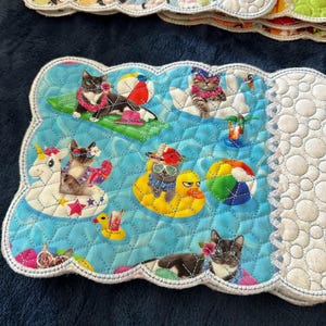 May include: A blue quilted placemat with a scalloped edge features a summer theme with cats wearing sunglasses, leis, and riding on inflatables. The placemat has a white border and a quilted pattern.