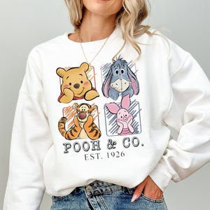 以下が含まれることがあります： 白のスウェットシャツ。くまのプーさん、イーヨー、ティガー、ピグレットの4人のキャラクターが描かれています。「POOH & CO. EST. 1926」の文字入り。カジュアルなスタイルです。