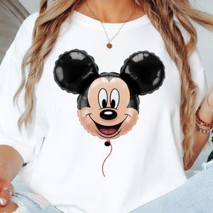 Puede incluir: Camiseta blanca con un dise&ntilde;o de globo de Mickey Mouse. El globo tiene orejas negras, una cara sonriente y una cuerda roja. La camiseta es de corte holgado.