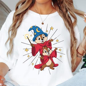 May include: White t-shirt featuring a cartoon illustration of two chipmunks dressed as wizards. The top chipmunk wears a blue wizard hat with a crescent moon and stars, and holds a wand. Both chipmunks wear red robes, surrounded by starbursts and yellow stars.