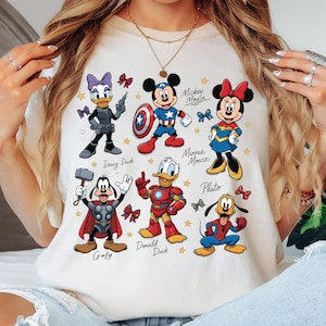 May include: Cream-colored t-shirt featuring cartoon characters Daisy Duck, Mickey Mouse, Minnie Mouse, Goofy, Donald Duck, and Pluto. Each character is depicted in a superhero or action-hero inspired outfit. The names of the characters are written below them.