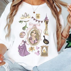 May include: White t-shirt featuring a collection of illustrations from a fairytale. The design includes a portrait, a tower, a frying pan, a crown, and other related elements in shades of pink, purple, and yellow.