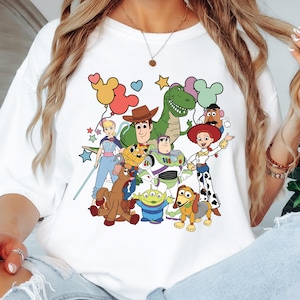 Op de afbeelding: Wit T-shirt met een kleurrijke afbeelding van personages uit de animatiefilm Toy Story. Het ontwerp toont Woody, Buzz Lightyear, Rex, Jessie en andere geliefde personages. Het shirt heeft een klassieke ronde hals.