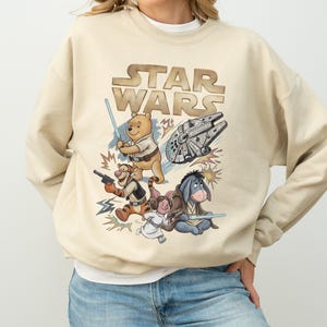 Puede incluir: Sudadera beige con un gráfico de "Star Wars" con personajes de Winnie the Pooh, el Halcón Milenario y sables de luz. El diseño incluye a Tigger, Eeyore y Piglet. El texto "STAR WARS" está en una fuente marrón estilizada.