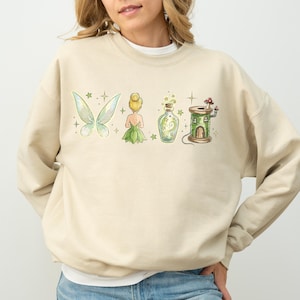May include: Beige sweatshirt with a watercolour design featuring fairy wings, a fairy, a potion bottle, and a mushroom house. The design is in shades of green, yellow, and beige. The sweatshirt is a casual, long-sleeved top.