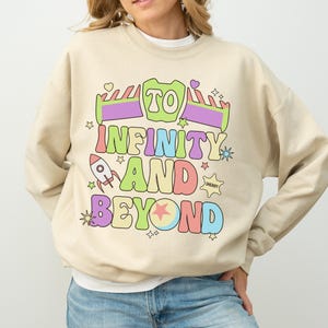 May include: Beige sweatshirt with the phrase "TO INFINITY AND BEYOND" in colorful, cartoon-style lettering. The design includes a rocket ship, a ball, and stars. The sweatshirt is paired with blue jeans.