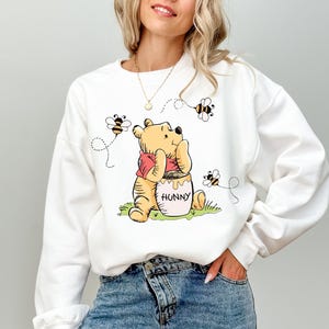 May include: White sweatshirt featuring a cartoon illustration of Winnie the Pooh with a honey pot and bees. The bear is wearing a red shirt. The word "HONNY" is on the honey pot. The bees have dotted flight paths.