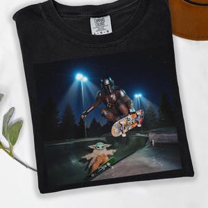 May include: Black Comfort Colors t-shirt featuring a graphic of The Mandalorian and Grogu skateboarding in a skate park at night. The image includes the text "COMFORT COLORS" on the upper right of the shirt.