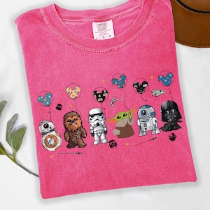 Puede incluir: Una camiseta rosa Comfort Colors con personajes de Star Wars: BB-8, Chewbacca, un Stormtrooper, Grogu, R2-D2 y Darth Vader. Sostienen globos con orejas de Mickey Mouse. La camiseta tiene cuello redondo.