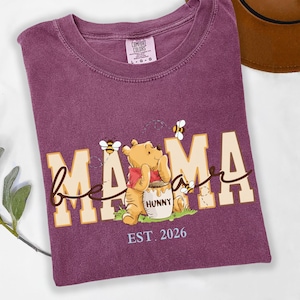 May include: A purple t-shirt with the words "bee mama" in a playful font, featuring a cartoon bear and bees. The bear is holding a honey pot. The text "EST. 2026" is below the graphic. The shirt is a Comfort Colors brand.