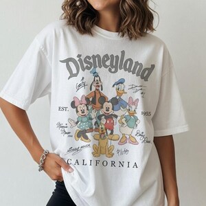 May include: White t-shirt with a Disneyland graphic featuring Mickey Mouse, Minnie Mouse, Donald Duck, and Goofy. The graphic includes the text "Disneyland California" and "Est. 1955". The shirt has short sleeves.