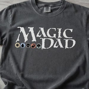 May include: A charcoal gray short-sleeve t-shirt featuring the white text MAGIC DAD printed on the chest. Below the text are five small circular icons representing sun, water drop, skull, flame, and tree symbols in various colors.