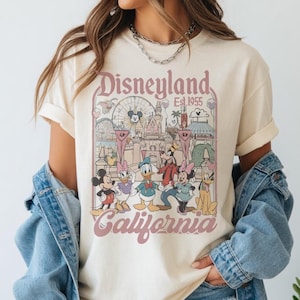 May include: Cream-coloured t-shirt with a Disneyland graphic featuring Mickey Mouse, Donald Duck, and other cartoon characters. The graphic includes the text "Disneyland Est. 1955 California" in a vintage style. The shirt is worn with a denim jacket.
