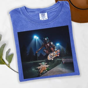 May include: Blue Comfort Colors t-shirt featuring a graphic of The Mandalorian and Grogu skateboarding in a skate park at night. The image shows the characters mid-air and on skateboards. The shirt has a crew neck and short sleeves.