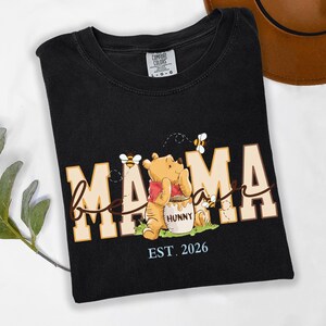 May include: Black t-shirt with the word "Mama" in a tan and white font, with a Winnie the Pooh graphic. The graphic includes bees and a honey pot. The text "EST. 2026" is below the graphic.