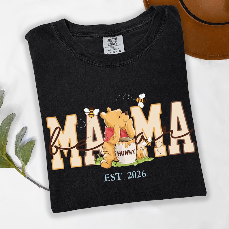 May include: Black t-shirt with the word "Mama" in a tan and white font, with a Winnie the Pooh graphic. The graphic includes bees and a honey pot. The text "EST. 2026" is below the graphic.