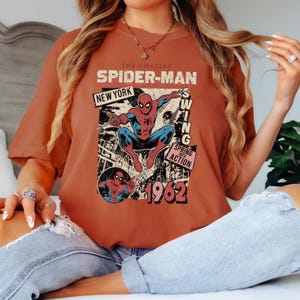 May include: Burnt orange t-shirt featuring a vintage Spider-Man graphic. The design includes the words "The Amazing Spider-Man," "New York," and "1962." The graphic shows Spider-Man in action, with a retro comic book style.