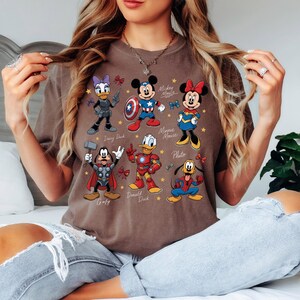 May include: A brown t-shirt featuring cartoon characters Daisy Duck, Mickey Mouse, Minnie Mouse, Goofy, Donald Duck, and Pluto, each in superhero-themed costumes. The names of the characters are printed below them. The shirt has a casual, relaxed fit.