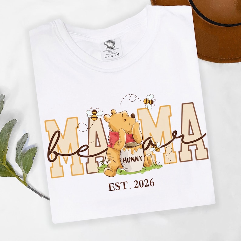 May include: White t-shirt with a graphic of Winnie the Pooh, the word "Mama" in tan block letters, and "bee" in cursive. Winnie the Pooh is holding a honey pot. The text "EST. 2026" is below the graphic.