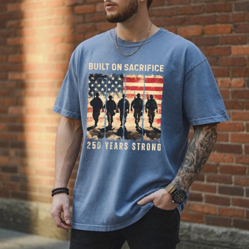 May include: Blue t-shirt featuring an American flag design with soldiers and the text "BUILT ON SACRIFICE" and "250 YEARS STRONG." The shirt has a crew neck and short sleeves. The shirt is made of a soft material.