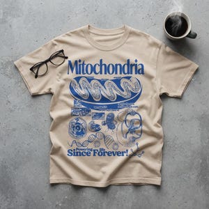 May include: A beige short-sleeve t-shirt featuring a blue graphic print of a mitochondrion, DNA strands, and scientific diagrams. The text reads Mitochondria, Powering life Since Forever!, and other small labels. Black eyeglasses rest on the fabric.