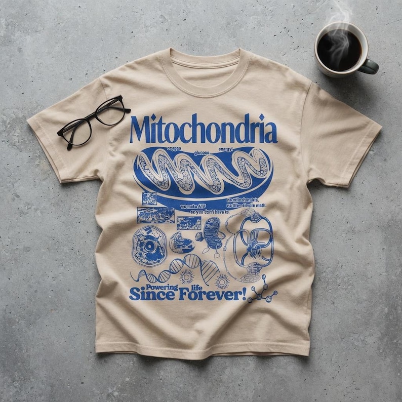 May include: A beige short-sleeve t-shirt featuring a blue graphic print of a mitochondrion, DNA strands, and scientific diagrams. The text reads Mitochondria, Powering life Since Forever!, and other small labels. Black eyeglasses rest on the fabric.