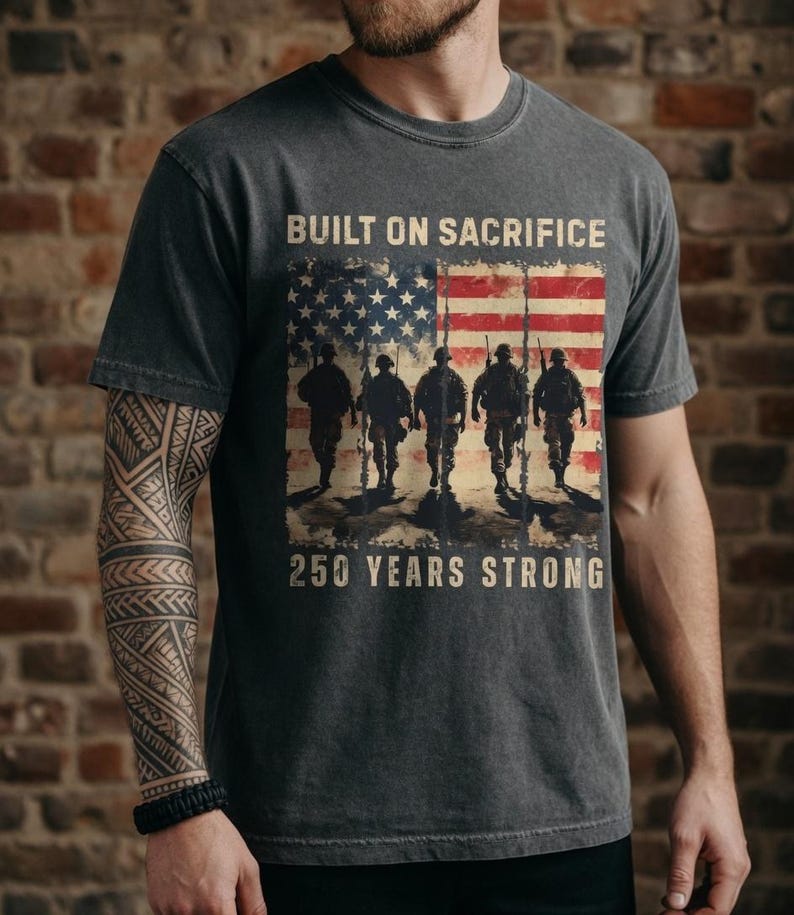 May include: Dark gray t-shirt featuring an American flag design with silhouettes of soldiers and the text "BUILT ON SACRIFICE" and "250 YEARS STRONG". The shirt has a vintage, distressed look.