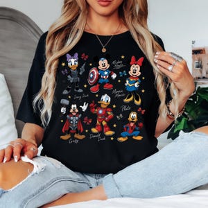 May include: Black t-shirt featuring cartoon characters Daisy Duck, Mickey Mouse, Minnie Mouse, Goofy, Donald Duck, and Pluto. The characters are dressed as superheroes. The shirt has a crew neck and short sleeves.