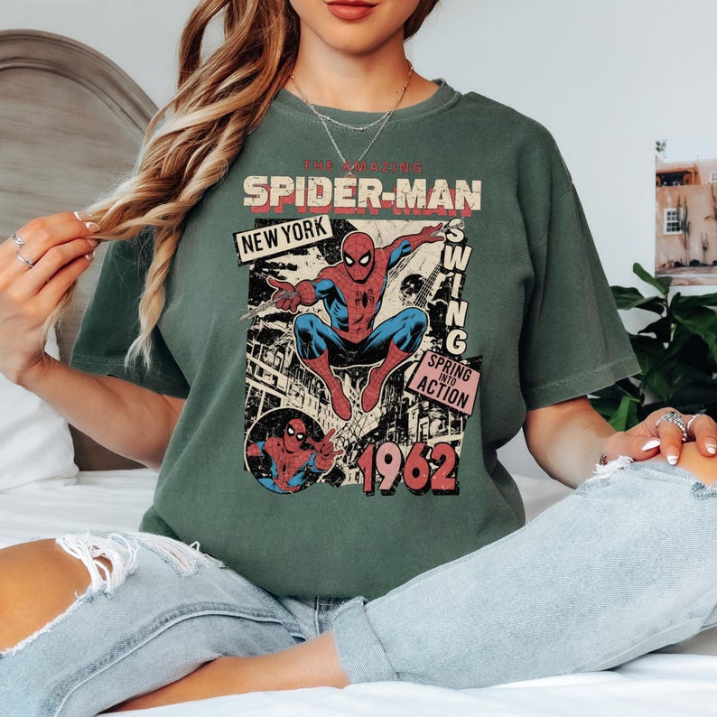 May include: Green t-shirt featuring a vintage comic book design of Spider-Man. The graphic includes the text "The Amazing Spider-Man" and "New York", with the year "1962" prominently displayed. The superhero is depicted in his classic red and blue suit.